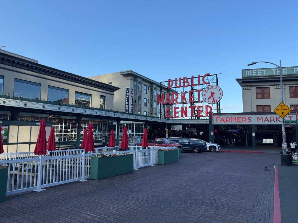 Exterior view of Pike Place Market in Seattle, showing the iconic Public Market Center sign and outdoor seating with red umbrellas.