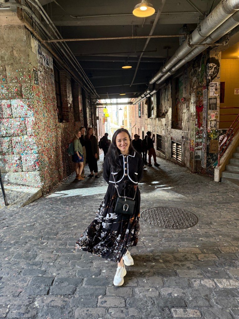 A person standing in a narrow alleyway in Seattle, surrounded by colorful gum on the walls, with other visitors visible in the background.