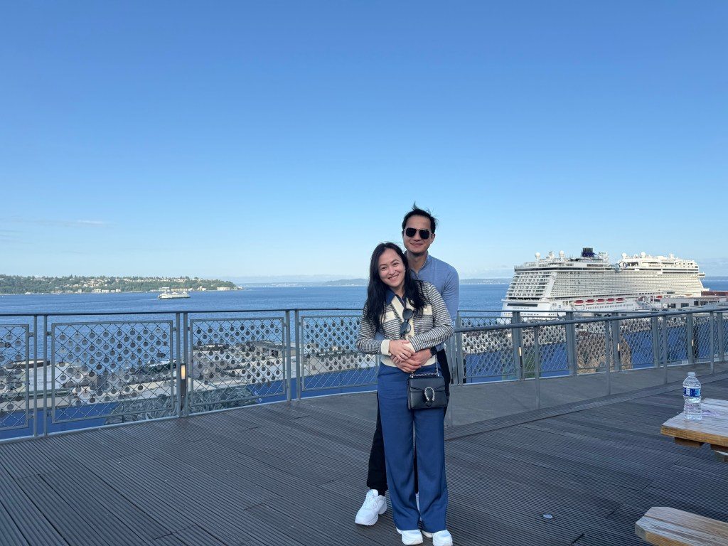 A couple posing together on a waterfront deck in Seattle, with a view of the ocean and a cruise ship in the background under a clear blue sky.